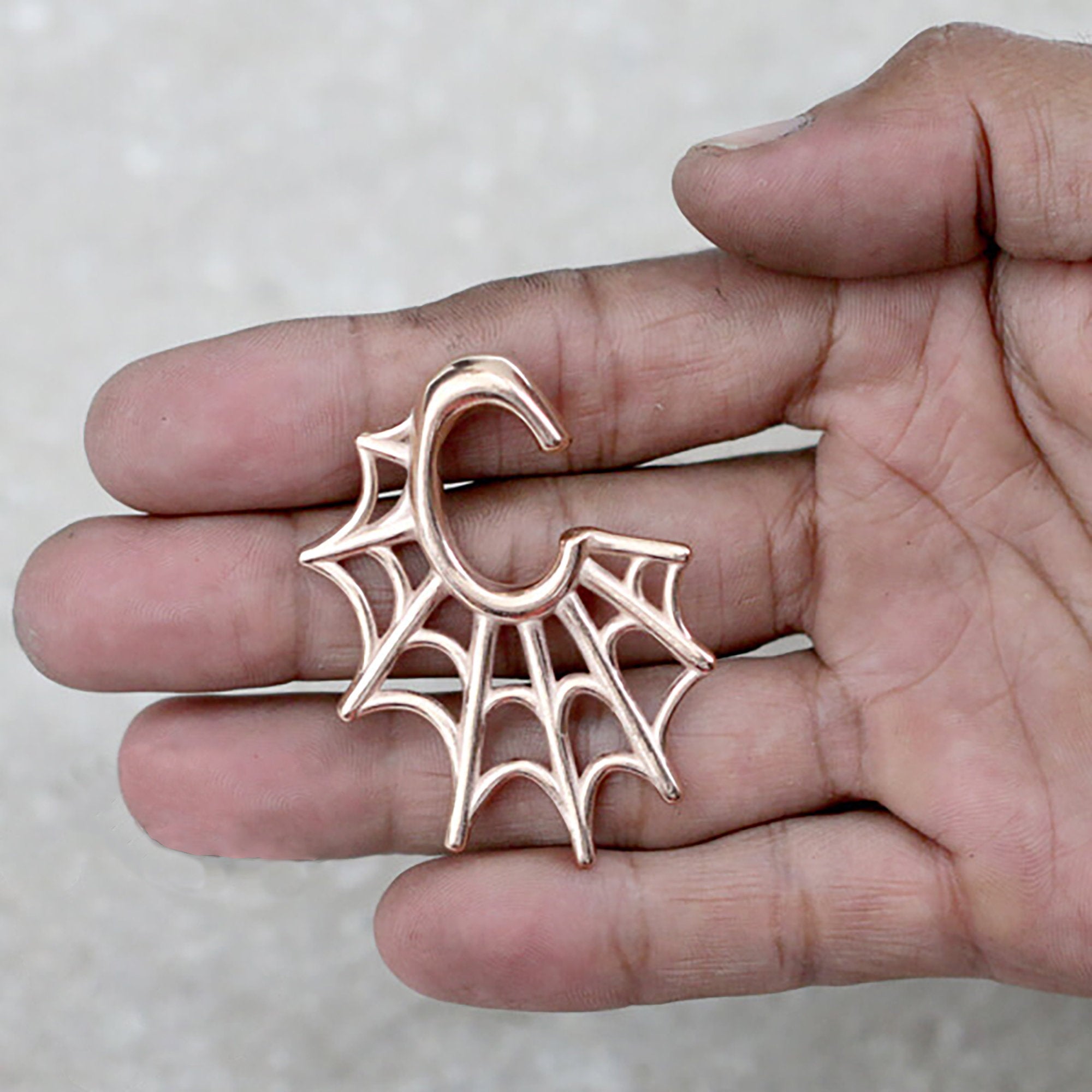 A hand holding a rose gold-colored, spider-web-shaped ear hanger. This stunning Spider Web Gothic Steel Ear Hanger in Rose Gold from Kaya Movement features a design resembling tribal ear weights. The earring forms a letter "C" at the top, with the intricate spider web pattern extending outward against a plain, light-colored background.